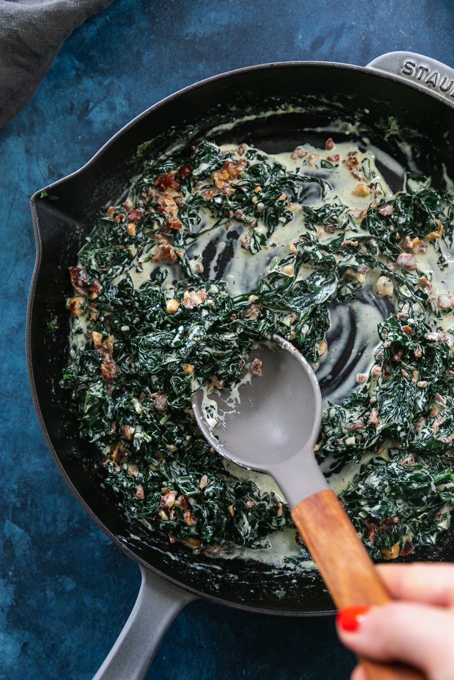 Overhead shot of a cast iron skillet filled with creamed kale and hazelnut bacon crunch and a spoon stirring it