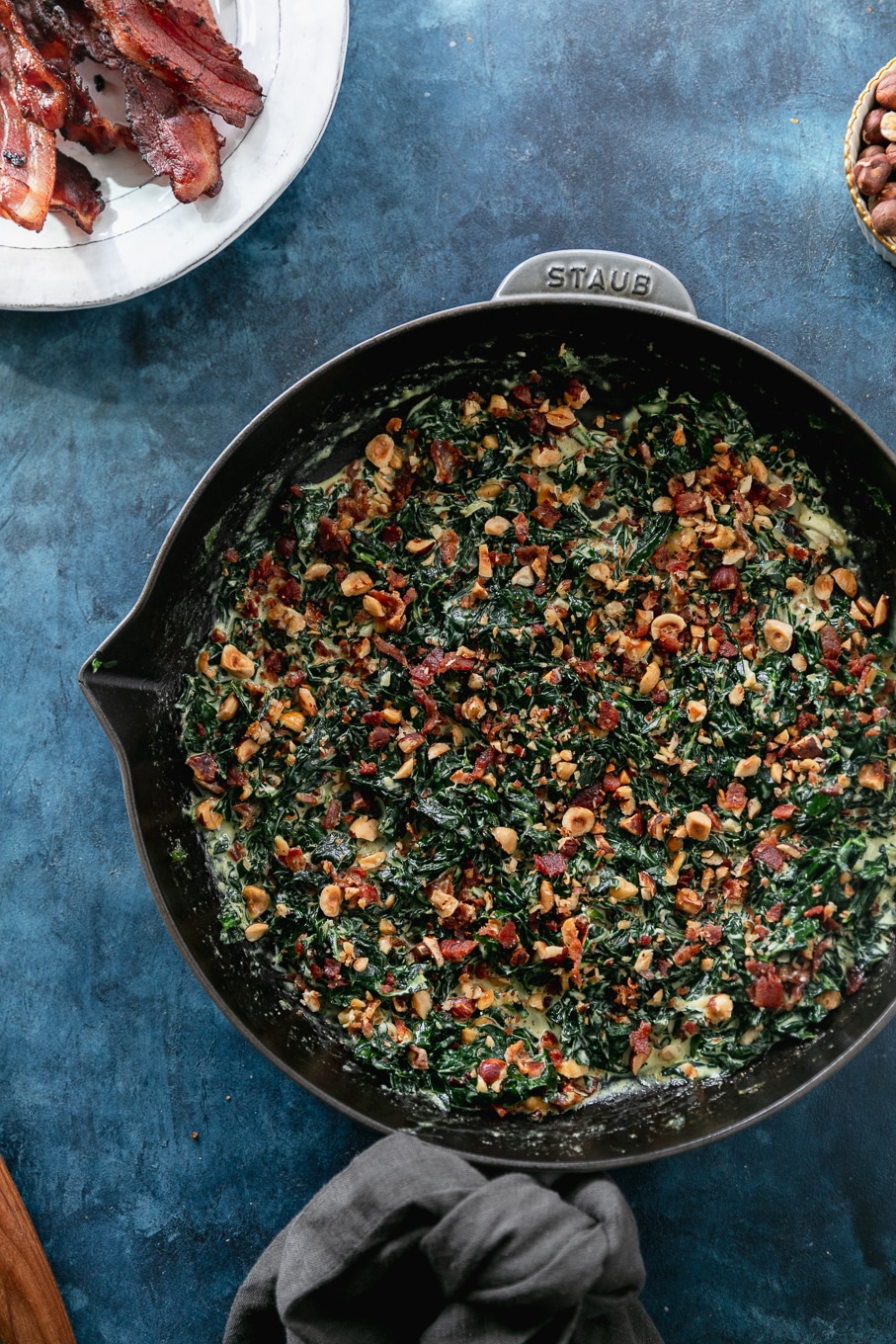 Overhead shot of a cast iron skillet filled with creamed kale and hazelnut bacon crunch with a plate of bacon in the upper left