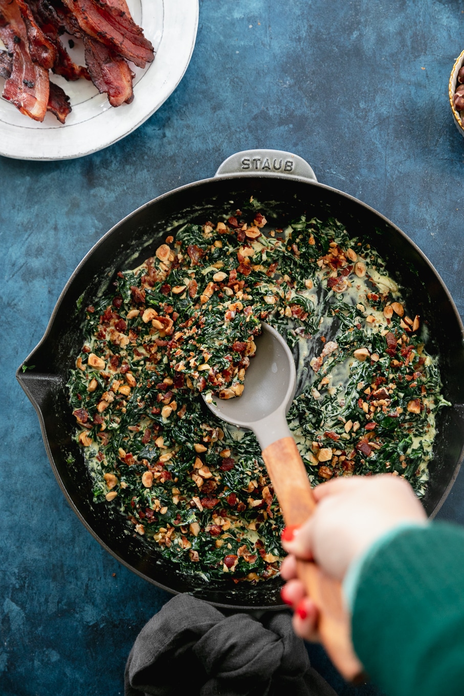 Overhead shot of a cast iron skillet filled with creamed kale and hazelnut bacon crunch and a spoon stirring it
