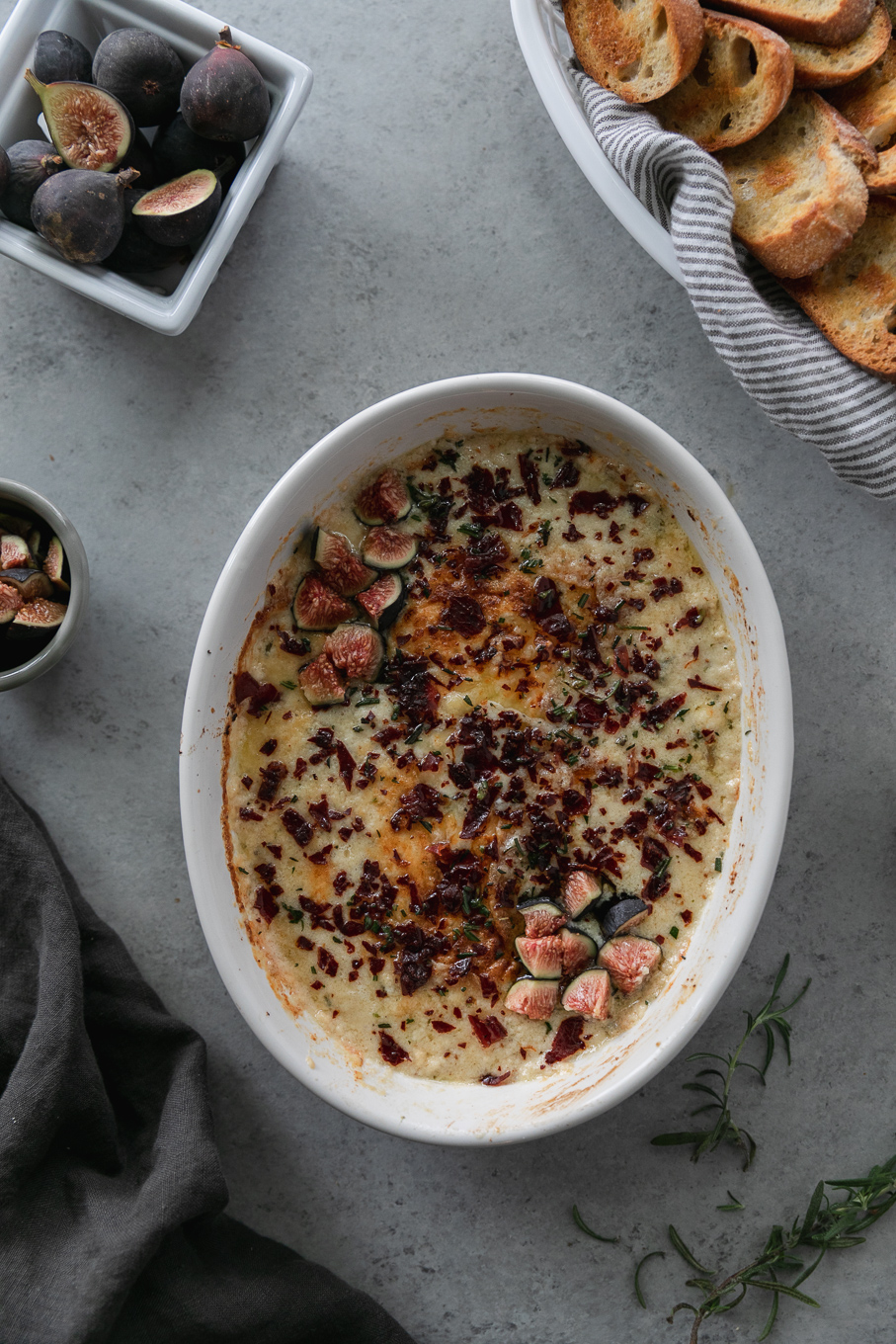 Overhead shot of a baking dish filled with a creamy dip topped with chopped figs and crispy prosciutto with a basket of crostini peeking out of the top right and a basket of figs in the top left