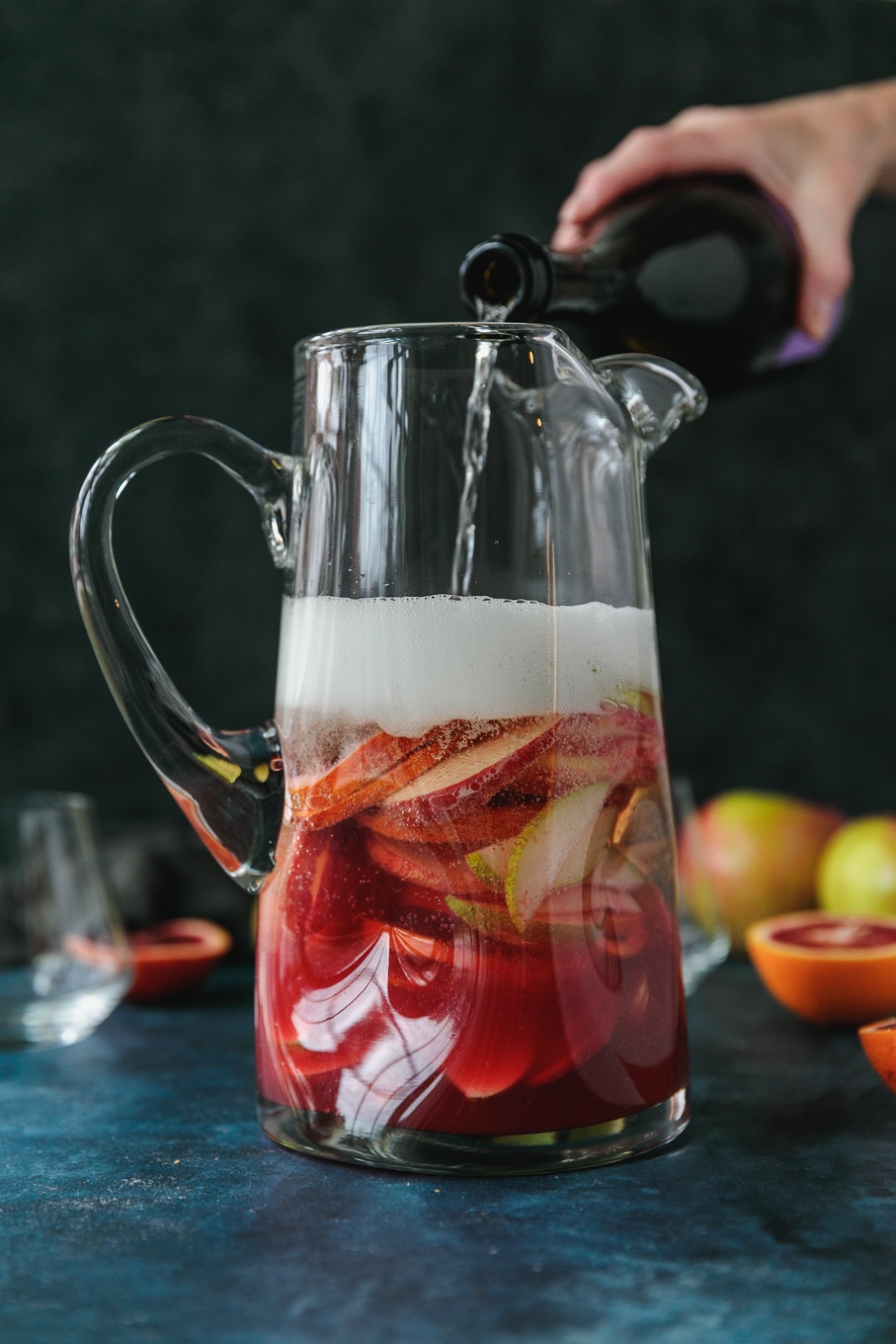 Forward facing shot of a pitcher filled with pink sparkling blood orange sangria filled with sliced apples, pears, and blood oranges with a bottle of champagne being poured into it