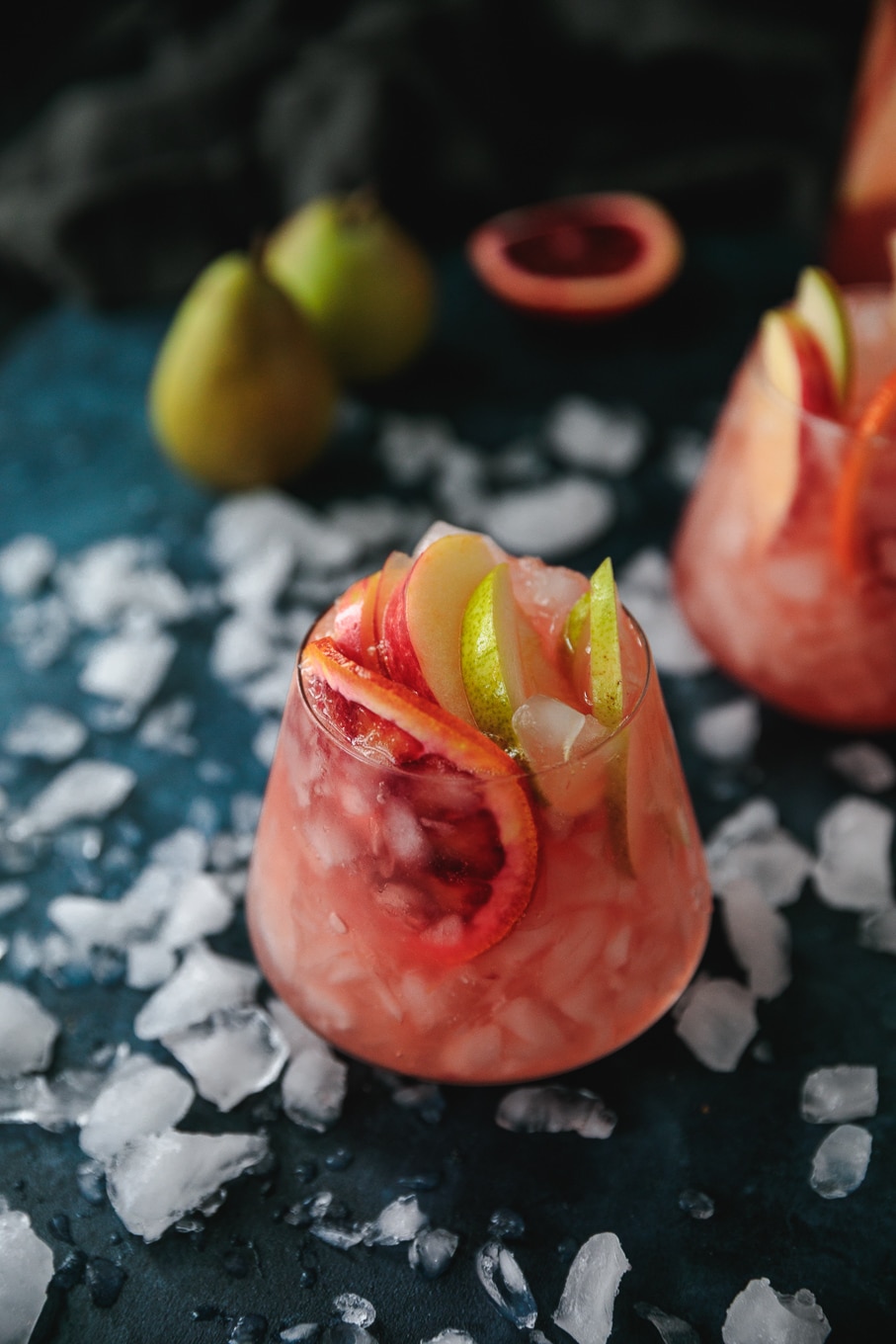 Close up shot of a glass of pink sparkling blood orange sangria with sliced apples, pears, and blood oranges sticking out against a blue background scattered with crushed ice