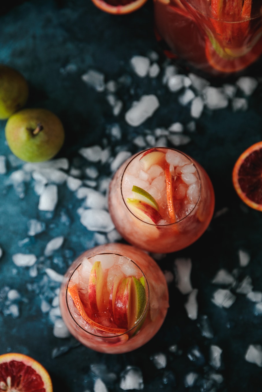 Overhead shot of two pink sparkling blood orange cocktails garnished with pears, apples, and blood orange slices, against a dark blue background with crushed ice scattered around