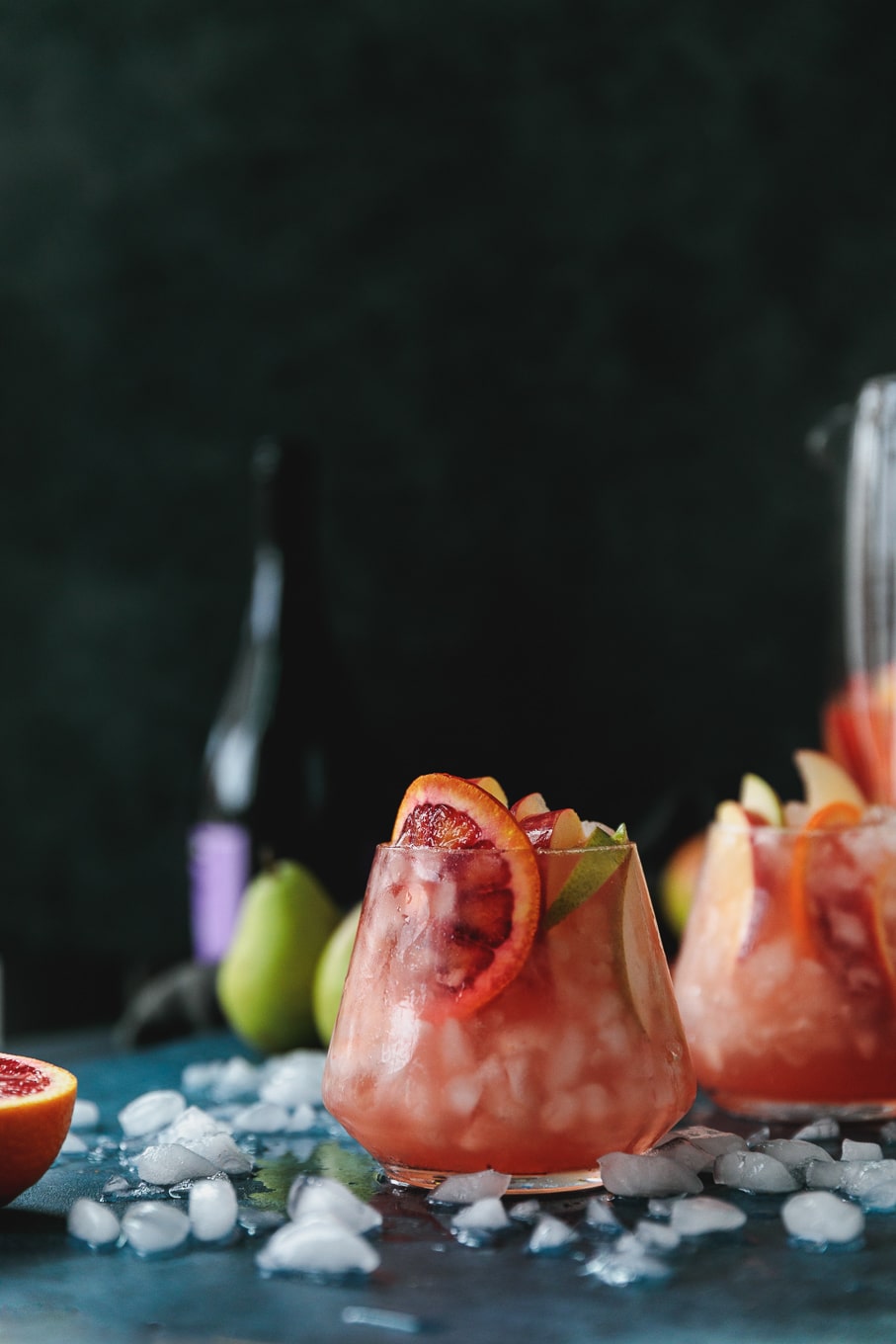 Forward facing shot of a glass of pink sparkling blood orange sangria garnished with a blood orange slice, with another cocktail behind it against a black background