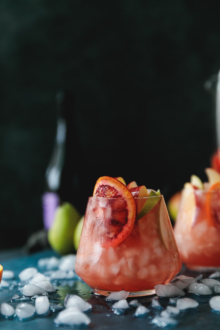Forward facing shot of a glass of pink sparkling blood orange sangria garnished with a blood orange slice, with another cocktail behind it against a black background