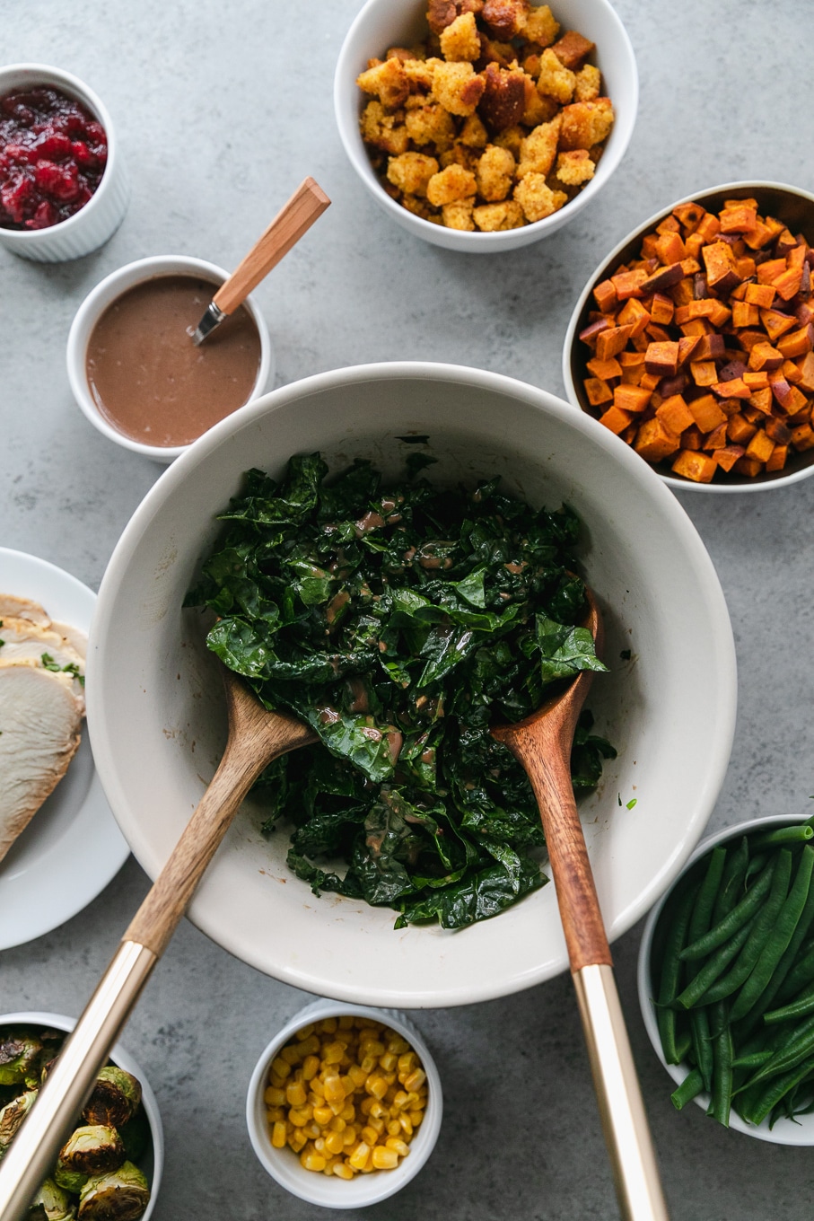 Overhead shot of a mixing bowl of kale with two wooden and gold salad tossers in the bowl, surrounded by a bowl of green beans, roasted sweet potatoes, a plate of turkey, and cranberry balsamic vinaigrette