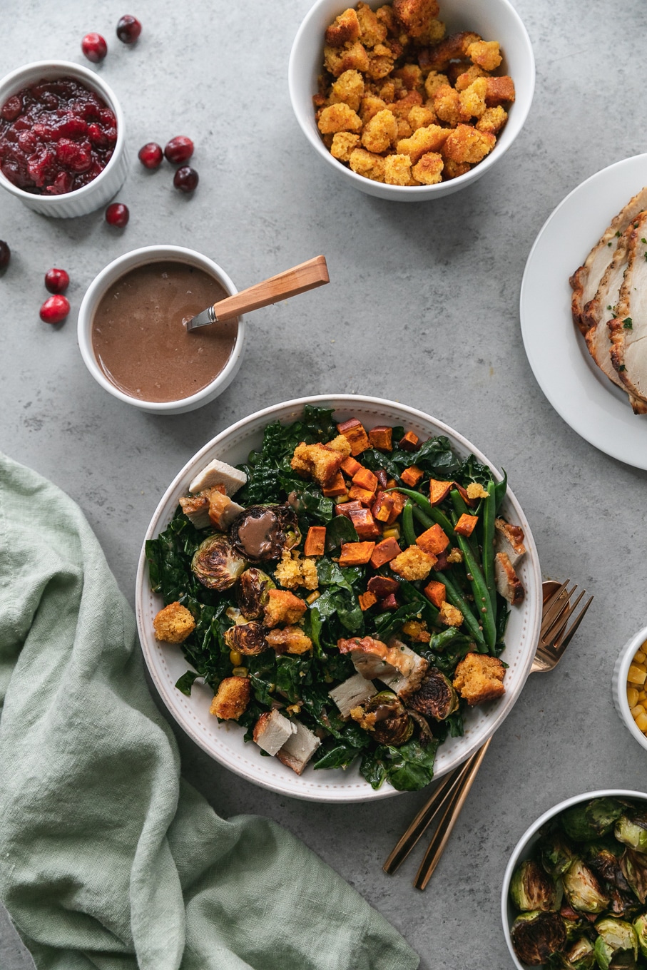 Overhead shot of a thanksgiving leftovers kale salad with roasted sweet potatoes, brussels, green beans, and cornbread croutons surrounded by a ramekin of vinaigrette, a plate of turkey, a ramekin of corn, and a bowl of brussels