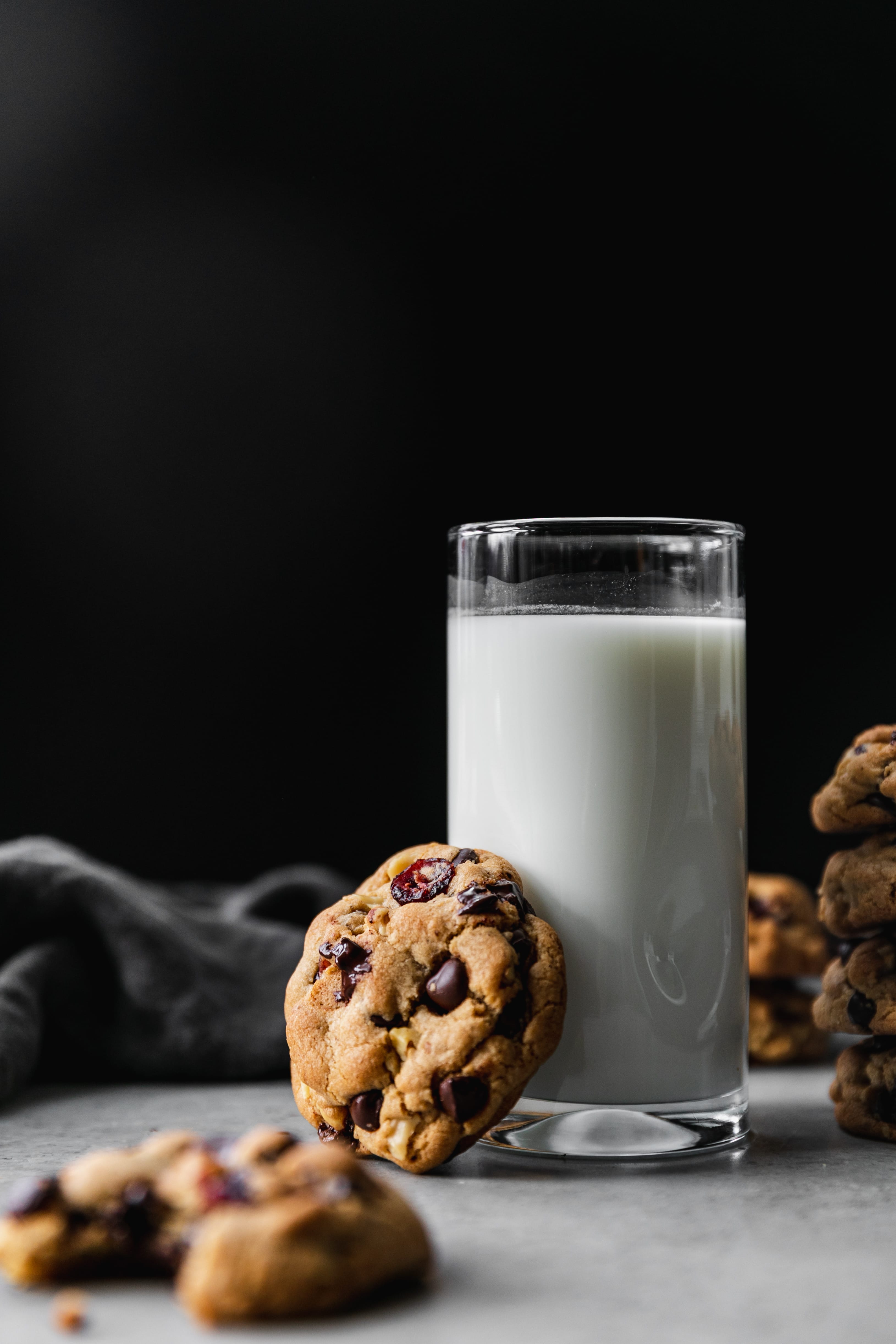Shot of a glass of milk against a black background with a dark chocolate cranberry walnut cookie resting up against it and a stack of cookies next to it