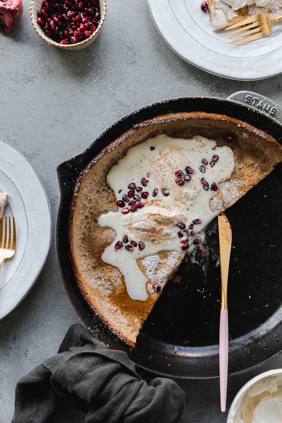 Overhead shot of a cast iron skillet with half of a dutch baby pancake in it, topped with melting whipped cream, pomegranate arils, and maple syrup, and a pink and gold knife resting in the skillet