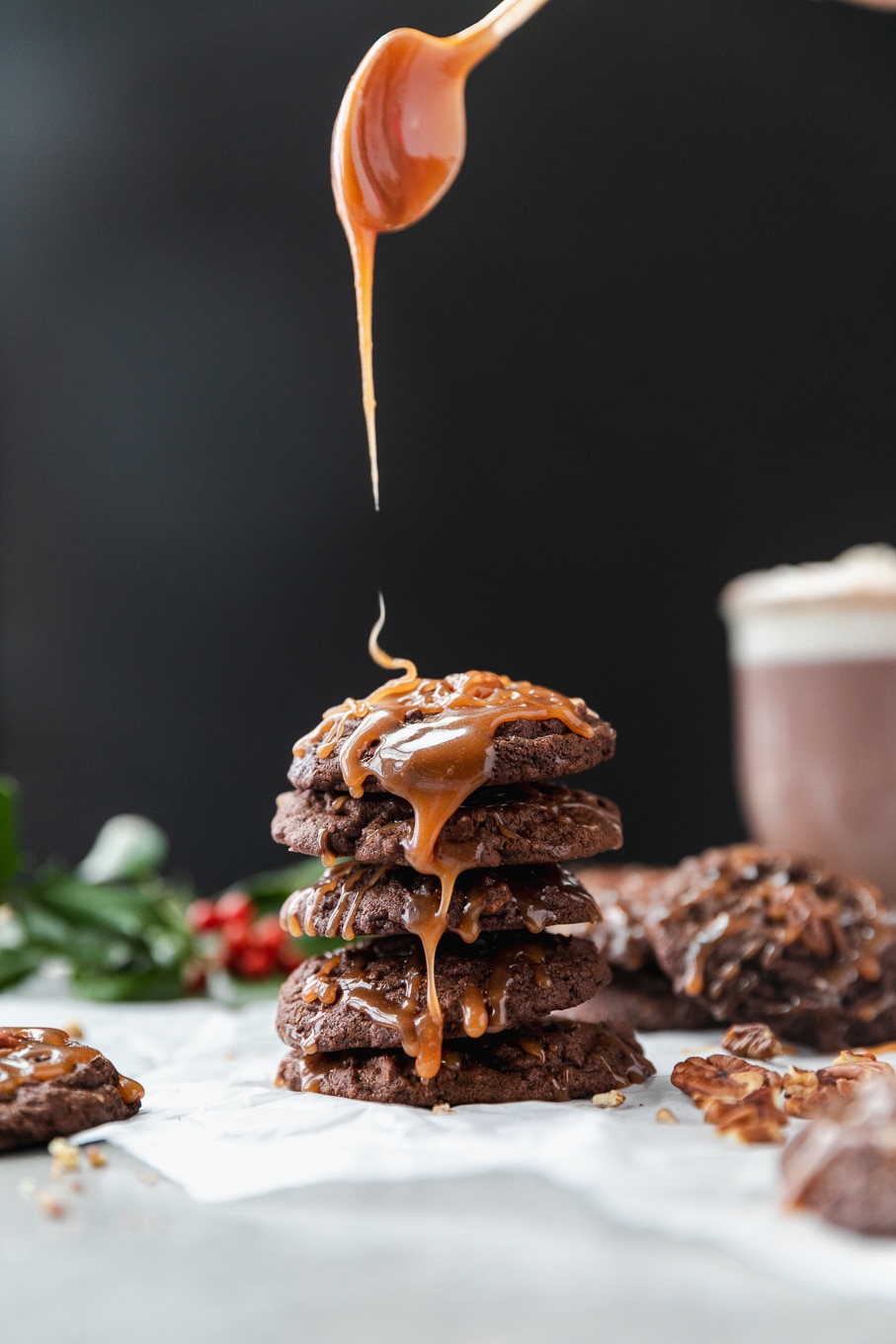 Forward facing shot of a stack of chocolate cookies being drizzled with salted caramel with a glass of hot chocolate against a black background with holly in the background