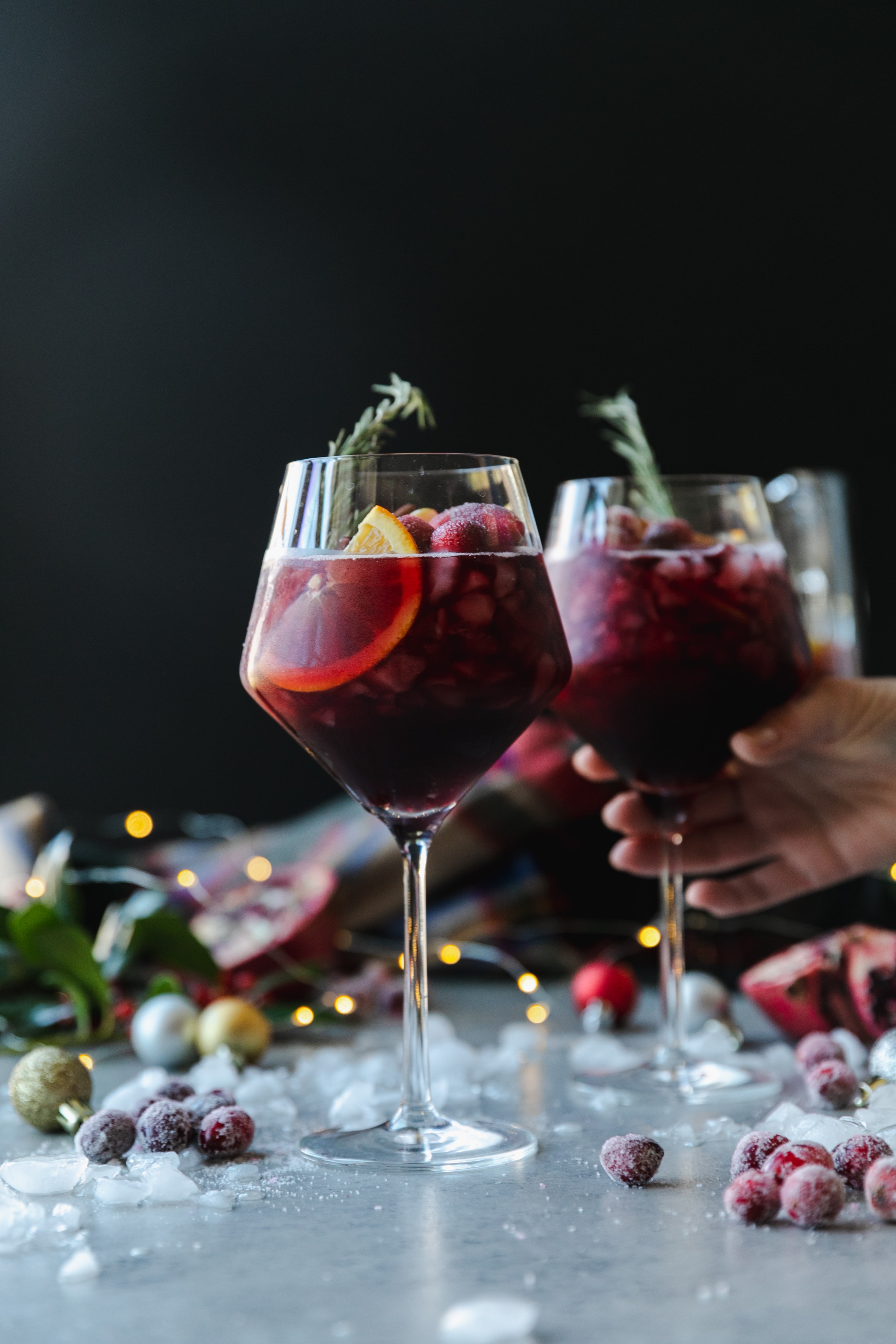 Forward facing shot of a wine glass filled with red sangria, garnished with a rosemary sprig, with another glass behind it with a hand reaching out to pick it up, with crushed ice, christmas ornaments, a plaid scarf, and twinkle lights in the background