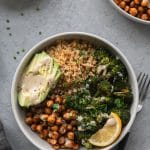 Overhead shot of a bowl with quinoa, chickpeas, roasted broccoli, sliced avocado, a lemon wedge, and tahini sauce with a black and silver fork next to the bowl