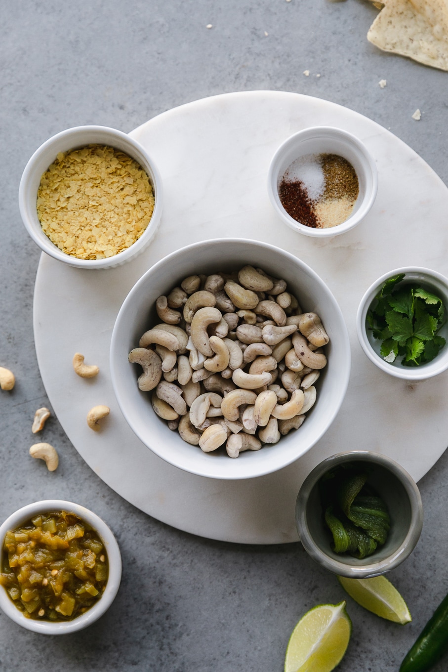 Overhead shot of a bowl of cashews surrounded by a bowl of nutritional yeast, a ramekin of spices, a ramekin of cilantro, a ramekin of charred jalapeño, and diced green chiles