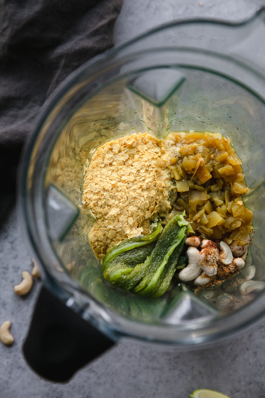 Overhead shot of a blender filled with nutritional yeast, diced green chiles, cashews, spices, and a charred jalapeño