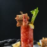 Forward facing shot of a bloody mary garnished with a grilled cheese sandwich on a skewer and a lobster claw sticking out of the drink with a lemon wedge and celery stalk with a grilled cheese sandwich next to it