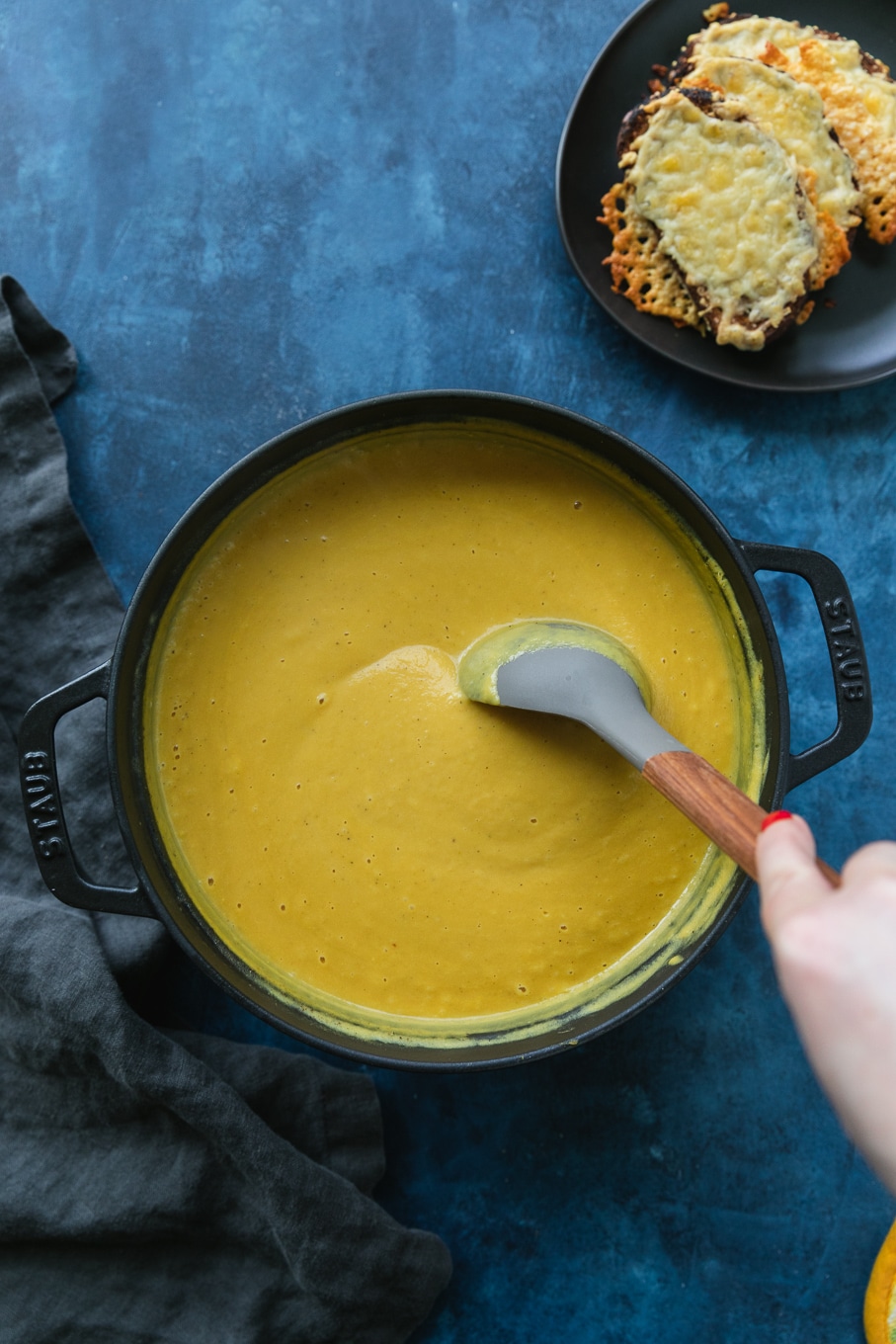 Overhead shot of a black cast iron pot filled with orange soup and a hand stirring it with a grey and wooden spoon
