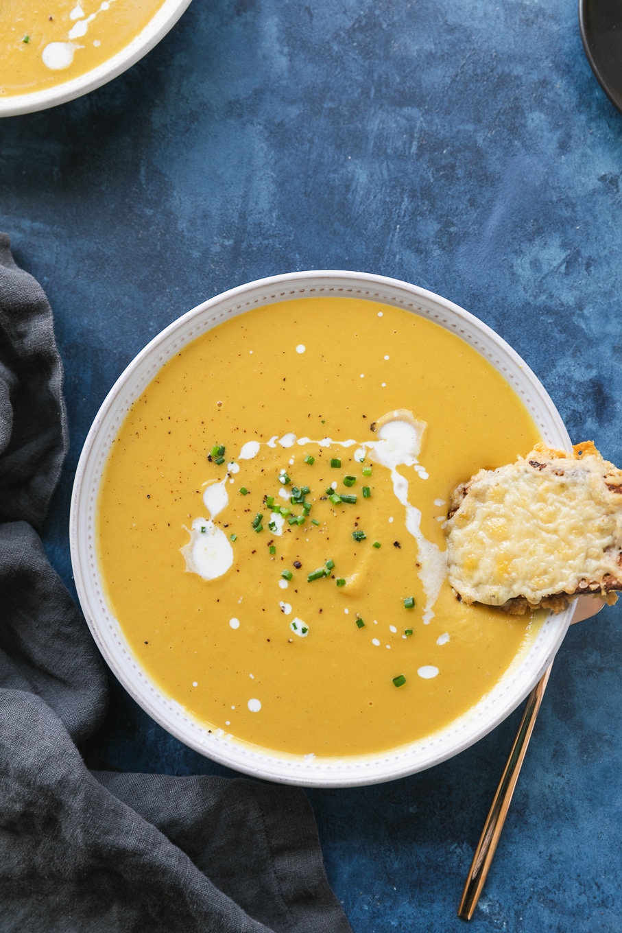 Overhead shot of a bowl of orange soup drizzled with cream and chives, with a cheesy toast resting in the soup