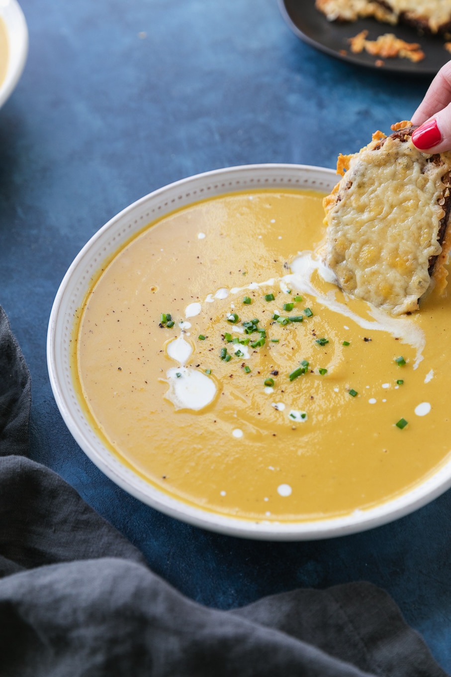 Close up shot of a bowl of orange soup with a hand dipping a cheesy toast in it