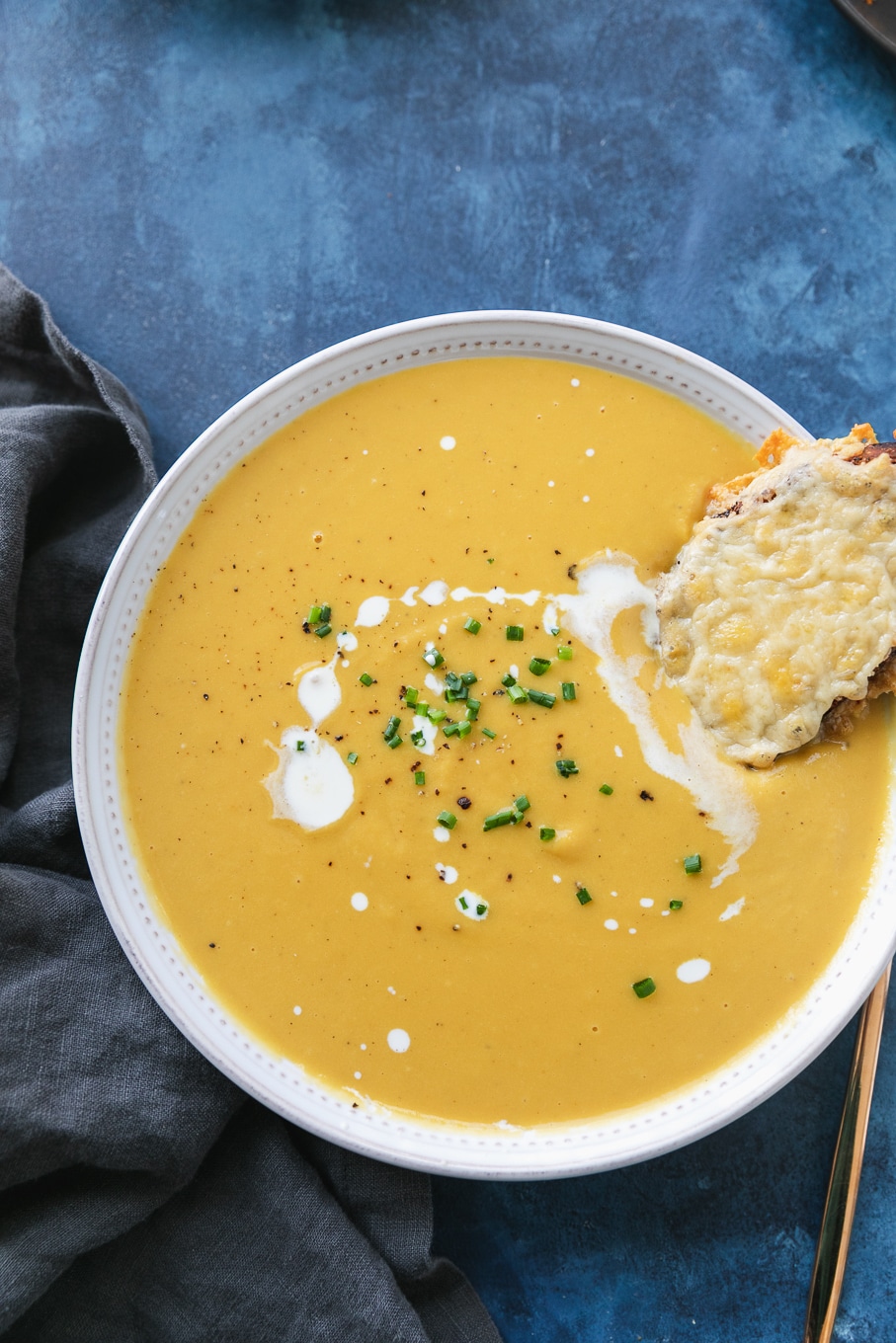 Overhead shot of a bowl of orange soup drizzled with cream and chives, with a cheesy toast resting in the soup