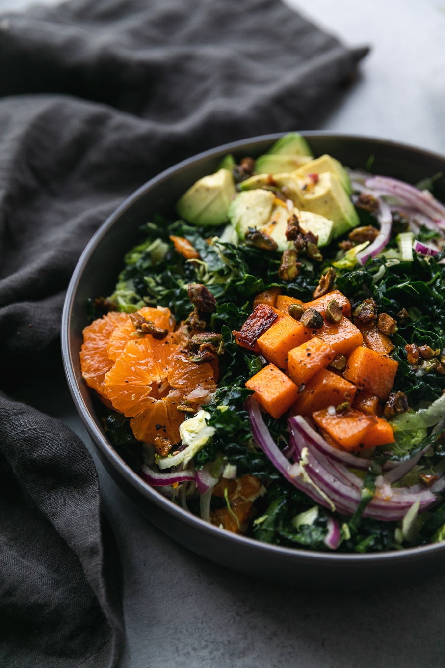 Close up shot of a kale salad with avocado, mandarins, red onion, butternut squash, and candied pistachios with a dark grey napkin next to it
