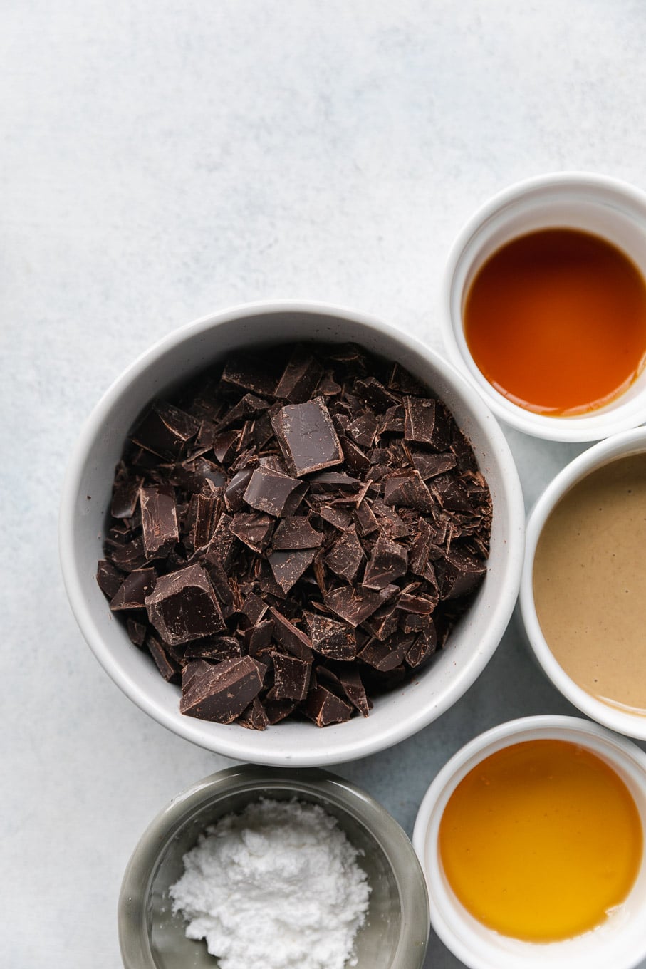 Overhead shot of a bowl of chopped dark chocolate, a bowl of tahini, a ramekin of honey, vanilla, and powdered sugar