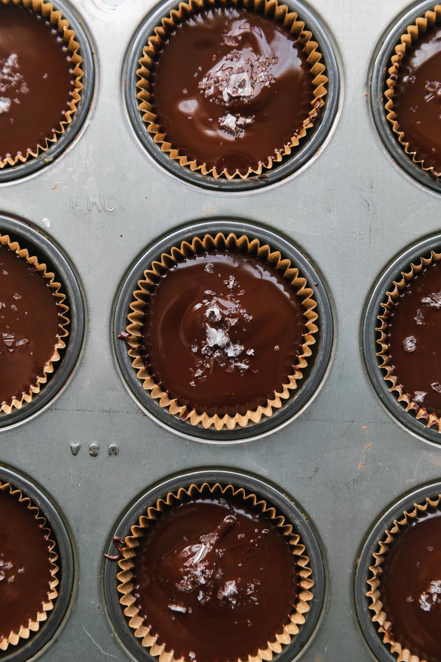 Overhead close up shot of a mini muffin tin filled with dark chocolate tahini cups and flaky sea salt on top of them