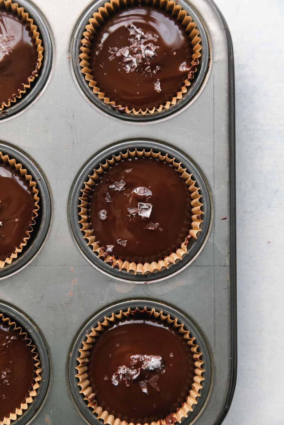 Overhead close up shot of a mini muffin tin filled with dark chocolate tahini cups and flaky sea salt on top of them