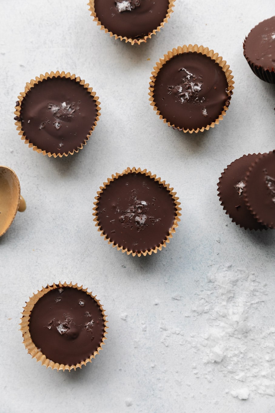 Overhead shot of dark chocolate tahini cups in parchment wrappers with a pile of flaky sea salt next to them