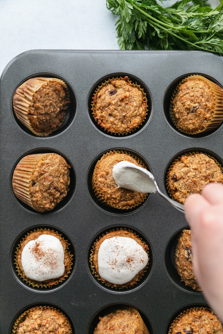 Overhead shot of carrot cake muffins in a muffin tin being topped with cream cheese glaze