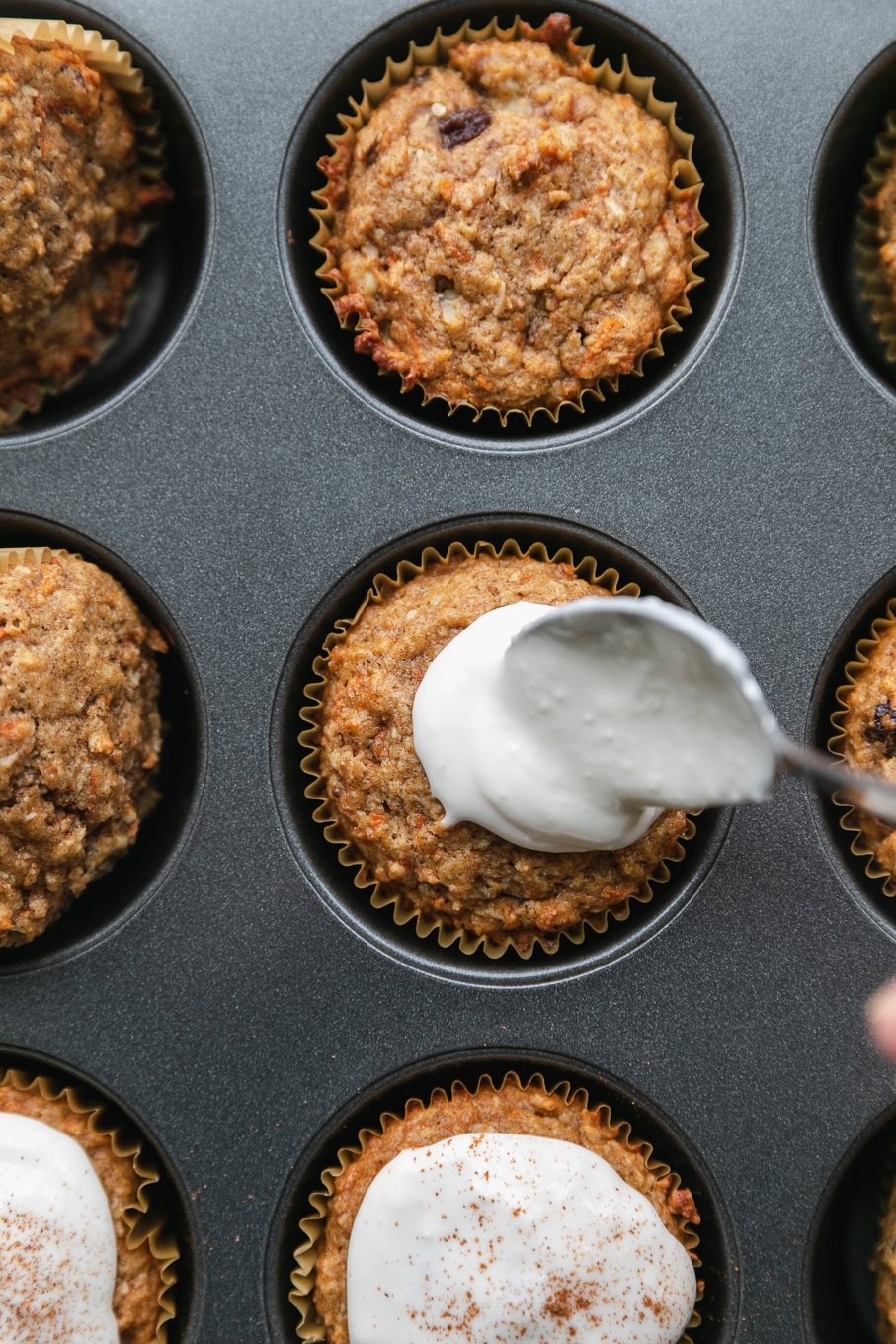 Overhead close up shot of a carrot cake muffin in a muffin tin being topped with cream cheese glaze