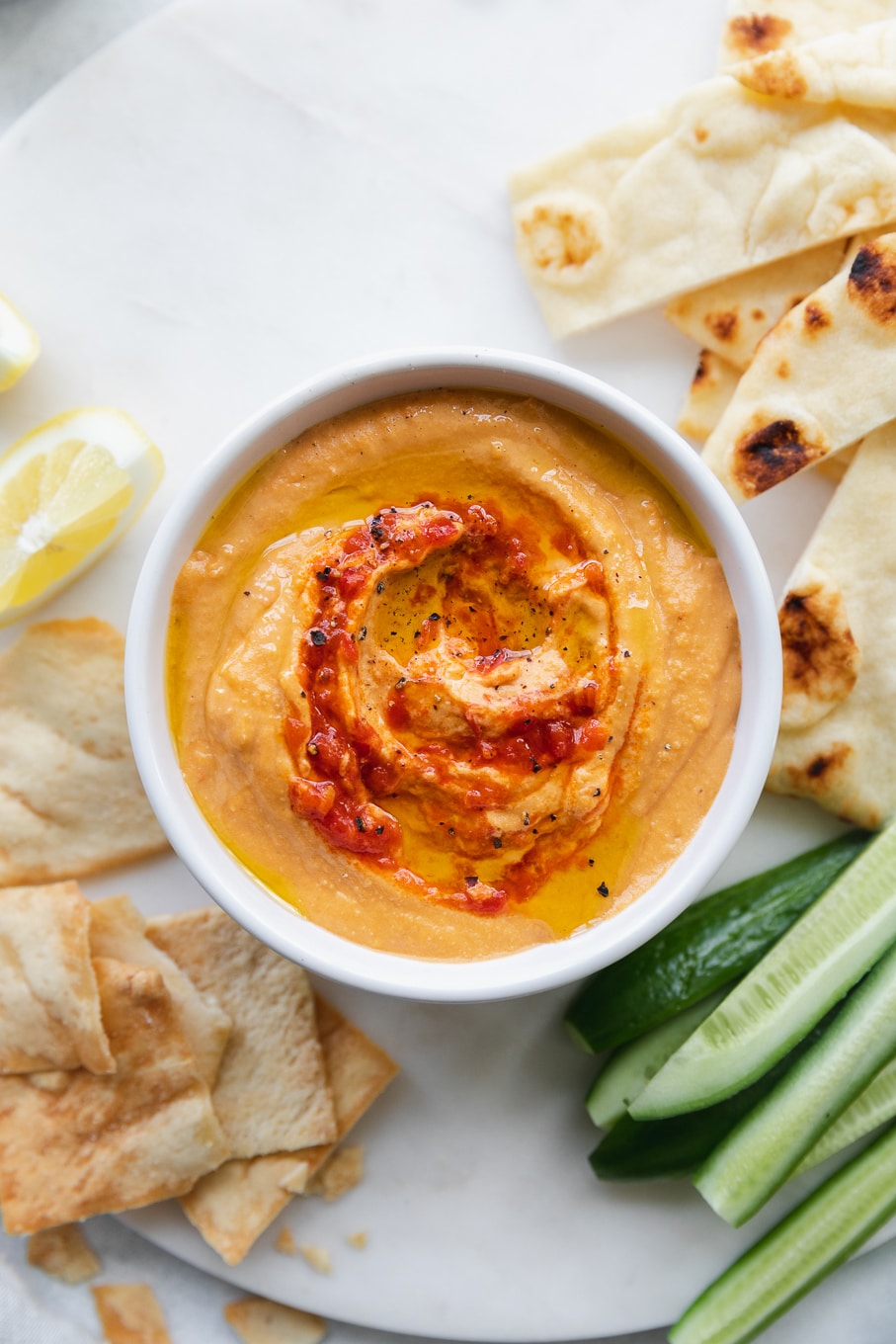Overhead shot of a bowl of harissa hummus surrounded by naan wedges, cucumber spears, and chips