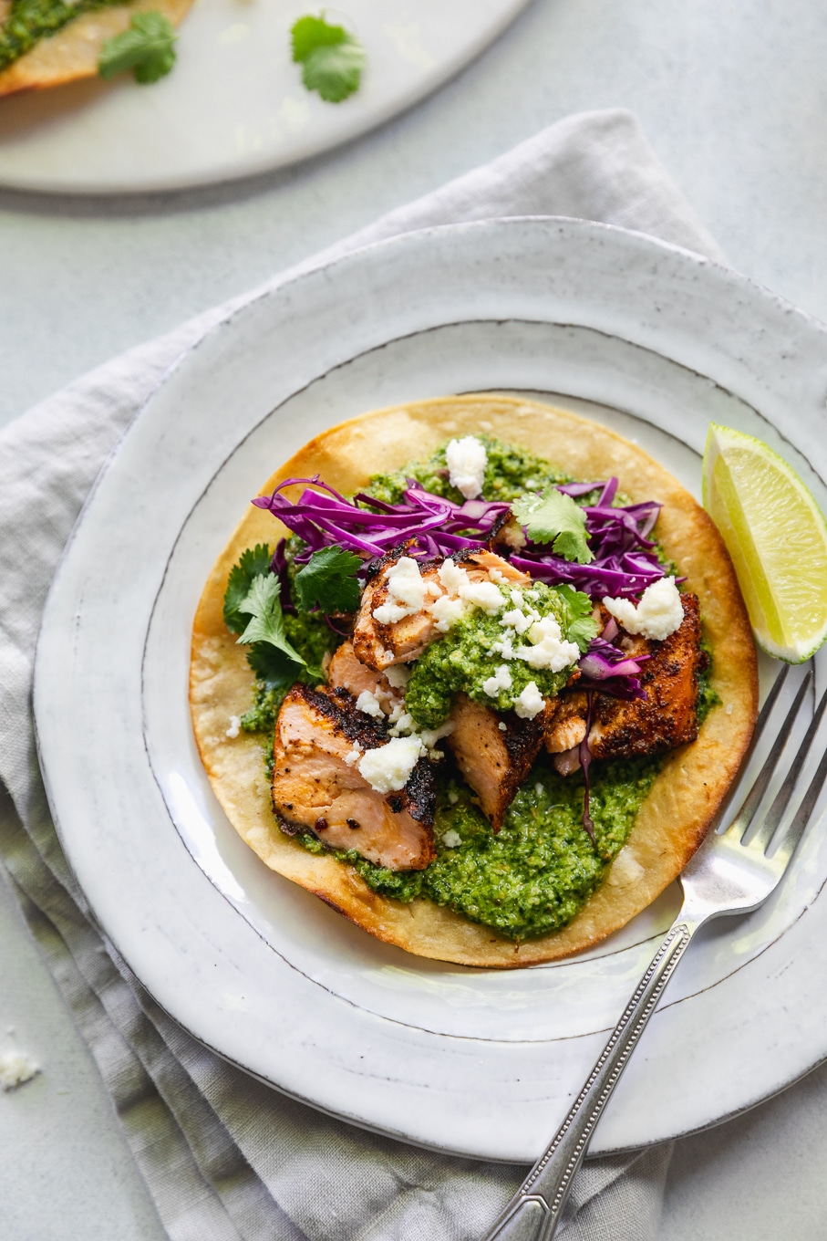 Overhead close up shot of a blackened salmon tostada with green sauce, red cabbage, queso fresco, cilantro, and a lime wedge on a white plate with a silver fork