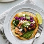 Overhead shot of a blackened salmon tostada with green sauce, red cabbage, queso fresco, cilantro, and a lime wedge on a white plate with a silver fork