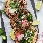 Overhead shot of 3 charred tortillas filled with grilled carne asada, chimichurri sauce, cilantro, sliced radishes, cotija cheese, and lime wedges