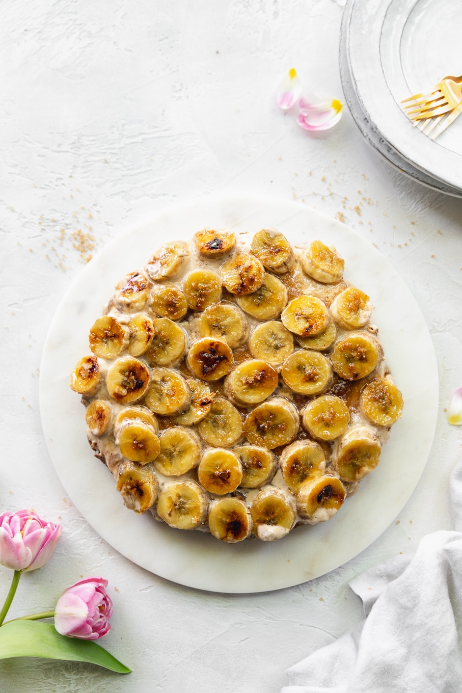 Overhead shot of a brûléed banana pie against a white background with pink tulips in the bottom left