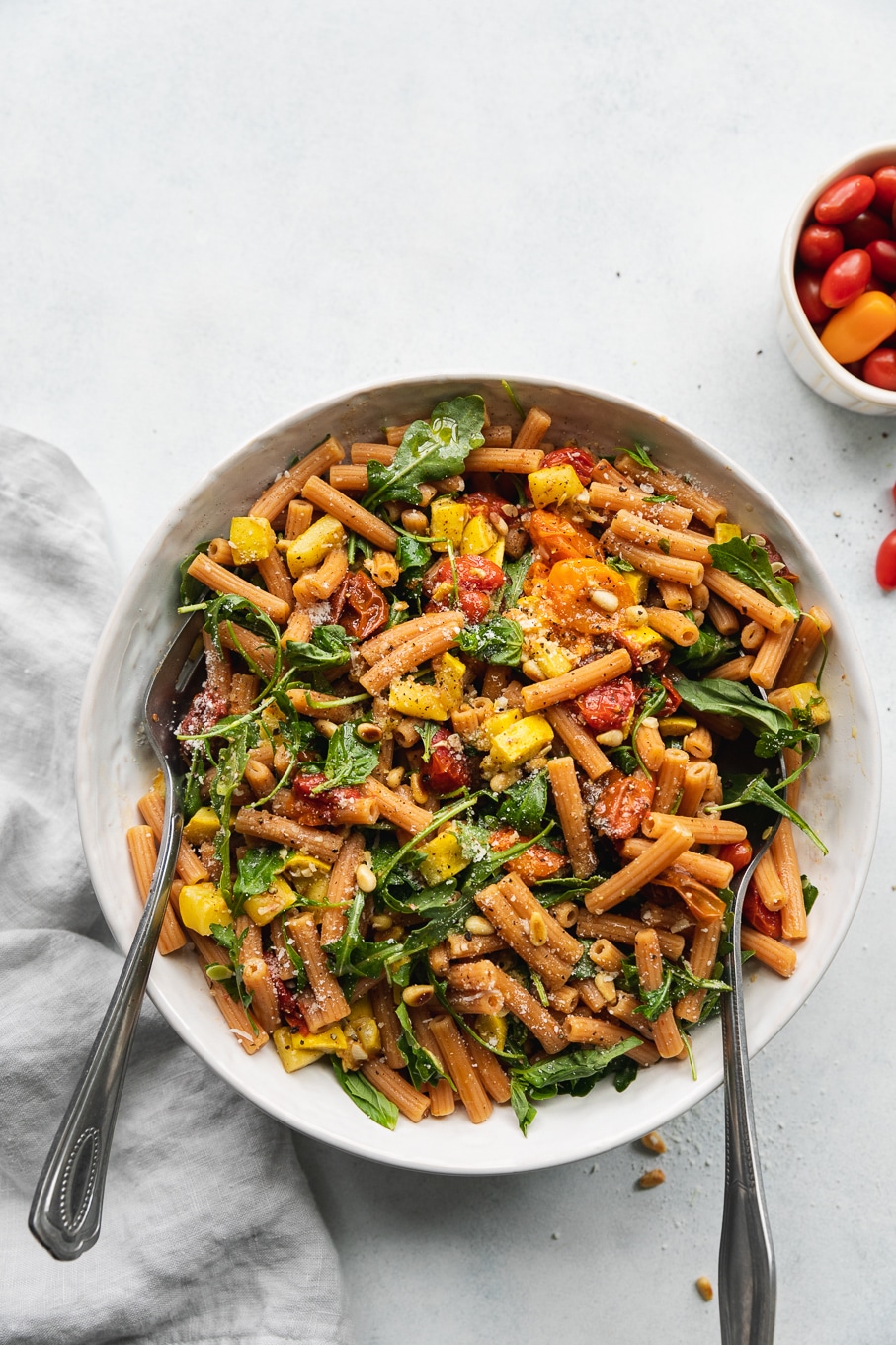 Overhead shot of a bowl of pasta filled with roasted tomatoes, summer squash, arugula, and parmesan cheese with two large silver utensils in the bowl