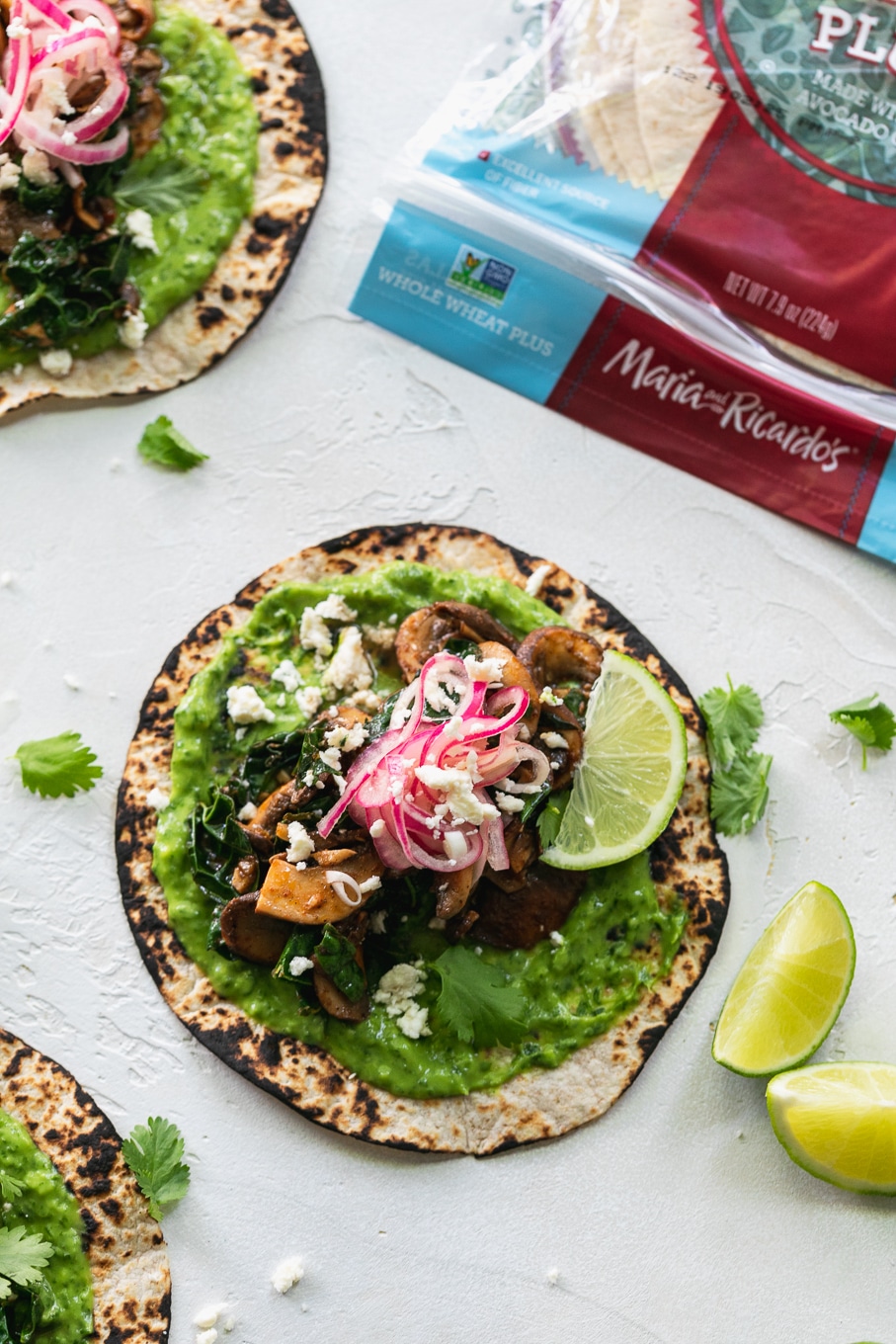 Overhead shot of a taco with green avocado sauce, pickled red onions, lime wedges, and a package of tortillas in the top right