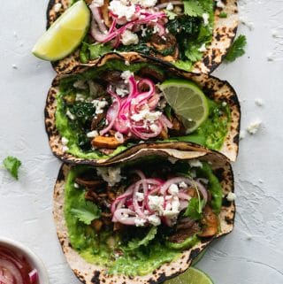 Overhead shot of 3 tacos with avocado cilantro sauce, pickled red onions, and lime wedges