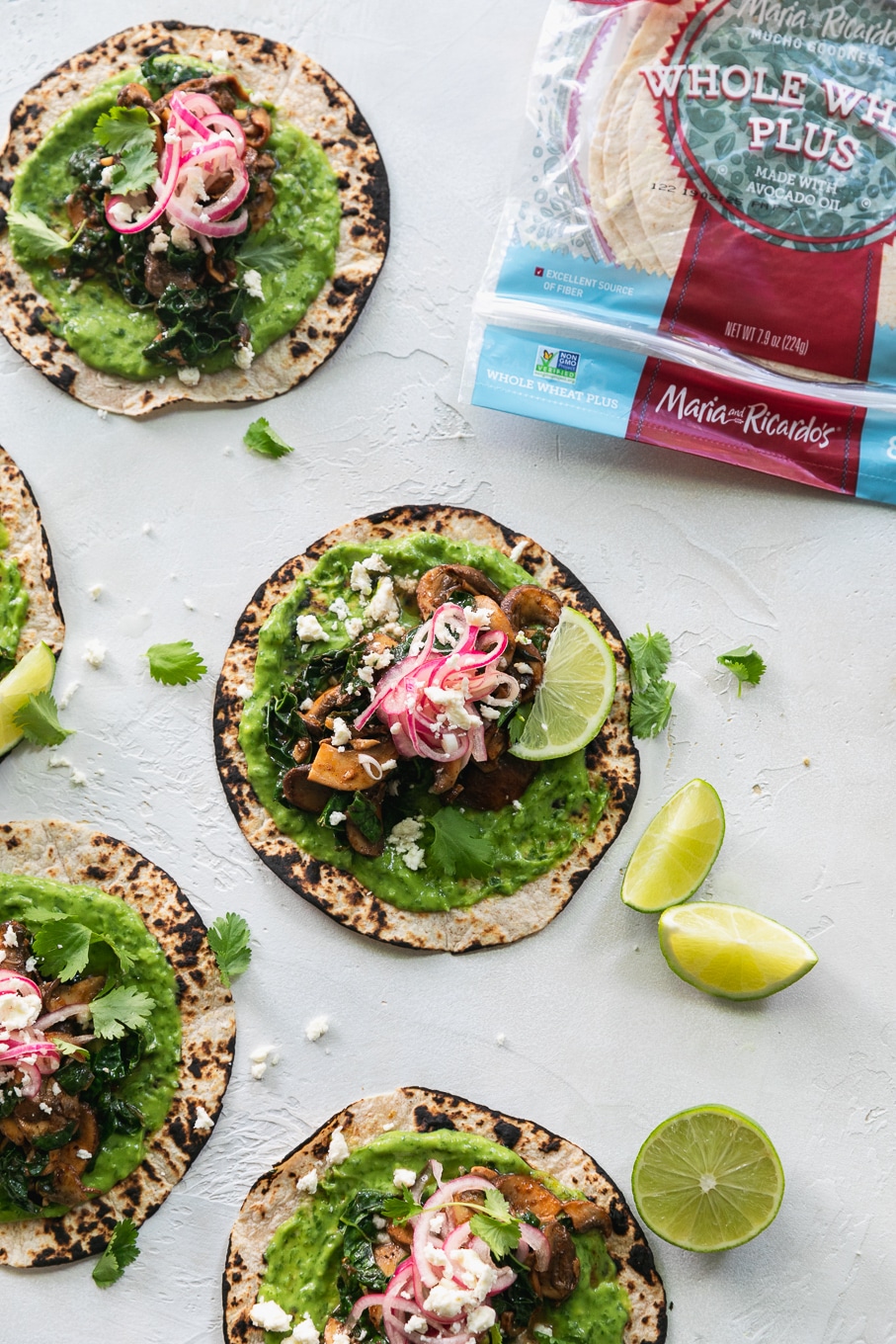 Overhead shot of tacos with green avocado sauce, pickled red onions, lime wedges, and a package of tortillas in the top right