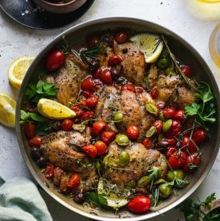 Overhead shot of a skillet filled with chicken, tomatoes, olives, herbs, and lemon